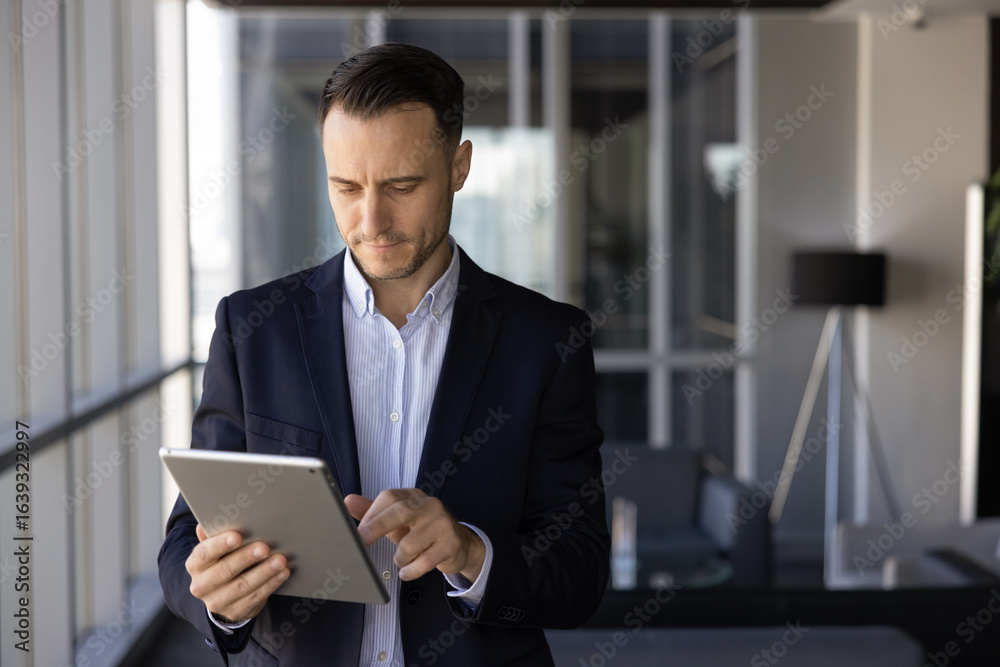 Focused busy Latin executive man using finance management application, trading service on tablet computer for business communication, typing on digital device, standing in office lobby Focused busy Latin executive man using finance management application, trading service on tablet computer for business communication, typing on digital device, standing in office lobby