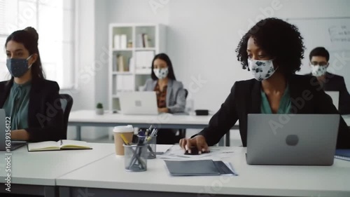 Two masked professionals work diligently at their desks in a bright environment, demonstrating workplace safety and productivity.