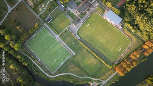 Aerial drone view of a sports complex with multiple football fields, running tracks, and green areas located along a canal.