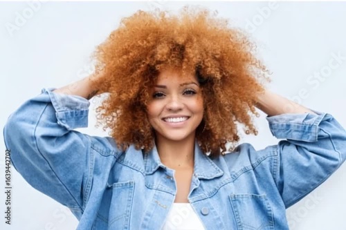 A beautiful woman with an afro hairstyle smiles while holding her hair in front of a white background, wearing a denim jacket and jeans.