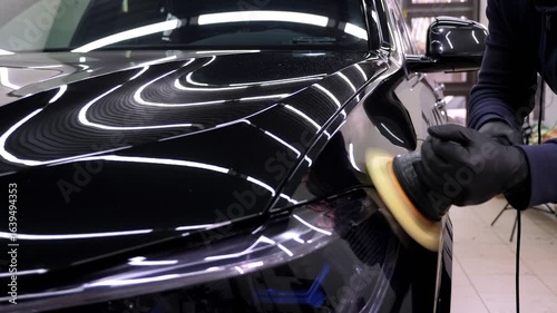 A close-up of a car mechanic polishing a black car with wax and a mechanical polishing machine. Modern car dealership and car body care.