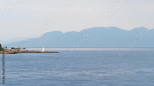 Wallpaper Mural Tranquil evening seascape of Aegina Island, Greece, filmed from the sea in summer. A white lighthouse stands on a rocky cape with trees, cars, and small buildings. Mountains fade into a hazy horizon  Torontodigital.ca