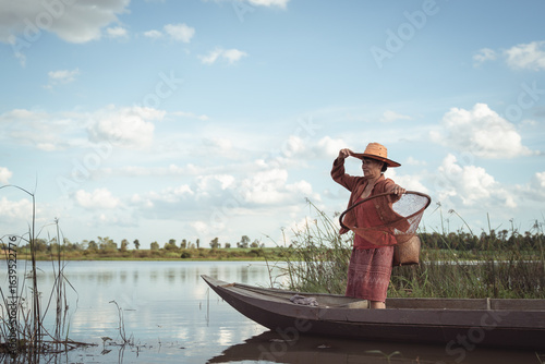Traditional Fisherwoman in a Boat, Casting a Net Amidst Serene Waters and Lush Greenery Under a Blue Sky with Fluffy White Clouds