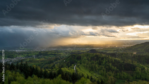 A dramatic and atmospheric view of a rain shower over the Murg Valley in the Northern Black Forest. Sun rays pierce through the dark clouds, illuminating the landscape below.