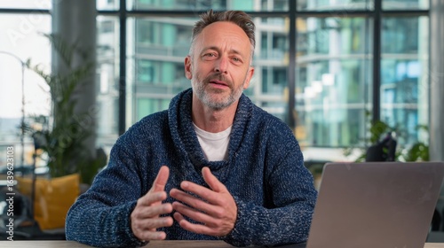 A mature man with a beard sits at a desk with a laptop, gesturing as he speaks, possibly during a video call or online presentation in a bright, modern setting.