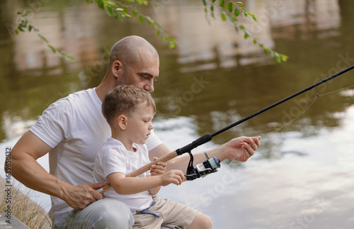 Father and young son fishing at calm lake in countryside at sunset. Horizontal family lifestyle photo with nature background and warm summer mood.

