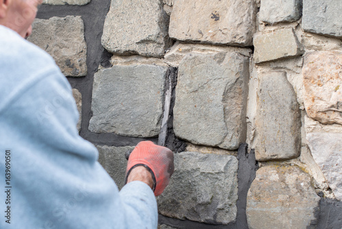 Builder applies mortar between stones in a wall using a long, narrow trowel during renovation work, demonstrating his masonry skills