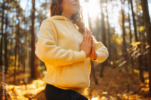 Young woman in bright sweater practicing yoga in autumn forest at sunset. Athletic woman exercising, stretching outdoors. Meditation, yoga and relaxation concept.