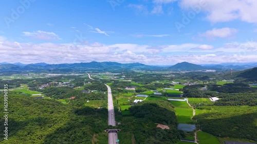 Aerial Drone View of Highway and Cars Driving Through Green Fields and Forest, with Vast Summer Landscape, Blue Sky, and Clouds for a Healing Scenery