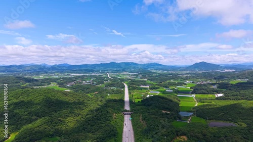 Aerial Drone View of Highway and Cars Driving Through Green Fields and Forest, with Vast Summer Landscape, Blue Sky, and Clouds for a Healing Scenery