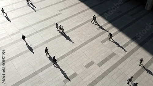 Highangle view of people with long shadows walking on a light gray paved plaza on a sunny day