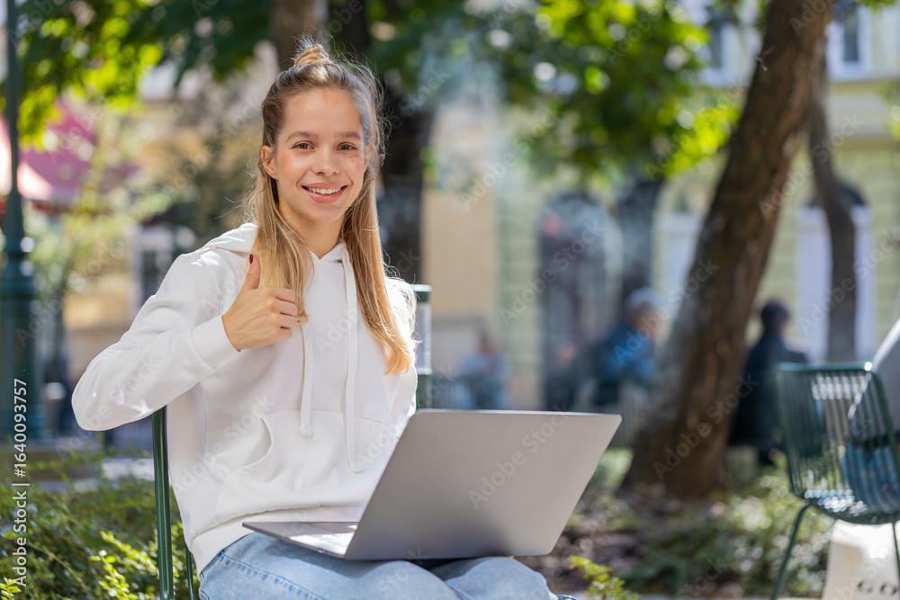 Happy young woman girl using laptop sitting on chair in urban city street outdoors. Caucasian girl working on notebook, sends messages, makes online purchases, watching movies and shows thumbs up. Happy young woman girl using laptop sitting on chair in urban city street outdoors. Caucasian girl working on notebook, sends messages, makes online purchases, watching movies and shows thumbs up.
