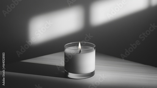 A simple white candle in a glass holder casts soft shadows on a surface, illuminated by natural light.