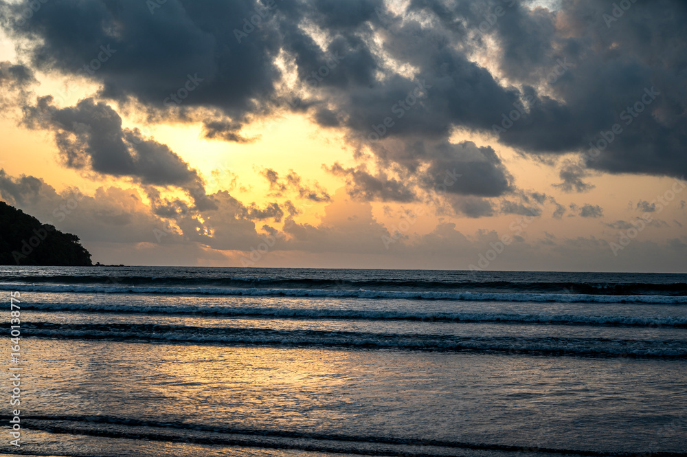 Fototapeta premium Sunset view of Vagator Beach Goa with Arabian Sea, golden yellow sky, dramatic grey monsoon clouds, and shimmering water reflecting the sun’s warm light in the evening.