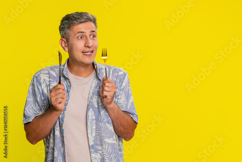 фотографія Middle-aged man holding knife and fork while looking directly at camera with eager hungry expression, showing desire for tasty meal
