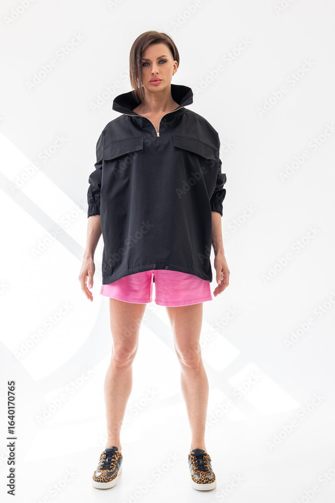 Fototapeta premium Elegant woman in black windbreaker jacket and pink shorts on light background. Fashionable female style. Studio photography. Vertical photo. High quality photo