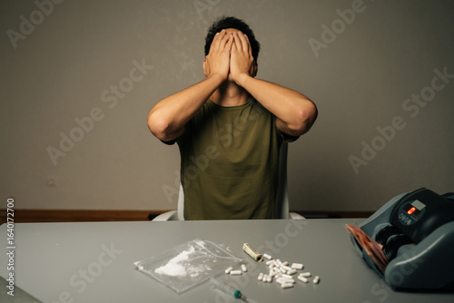 Studio shot of drug addiction struggle. Portrait of unrecognizable man covering face sitting at table with narcotics, banknotes, syringe, showing despair, dependency crisis. Concept of substance abuse