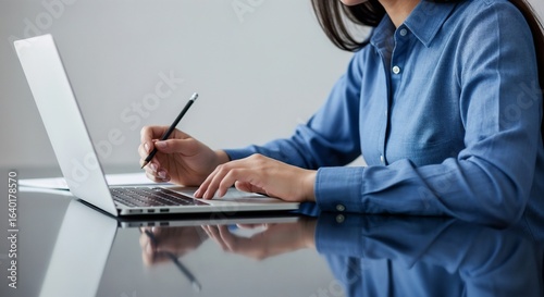 A woman in a blue shirt taking notes with a pencil next to a laptop on a reflective desk.