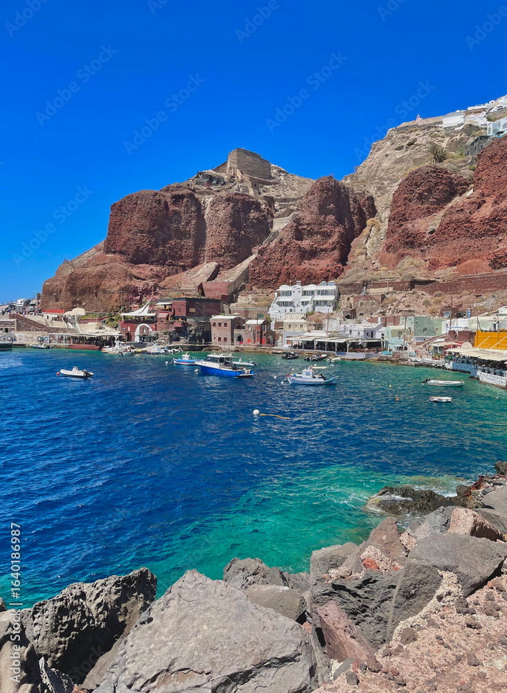 Obraz premium Picturesque harbor at the base of red volcanic cliffs, with boats floating on turquoise water in a charming Greek coastal village.