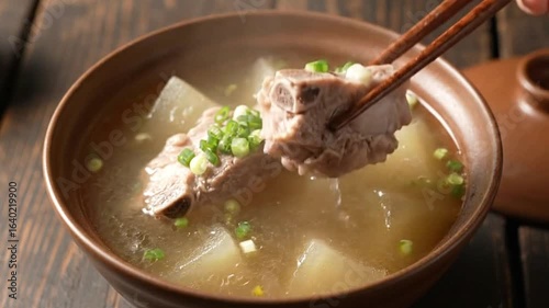 Steaming bone broth with ribs and daikon in a brown ceramic bowl lifted by chopsticks