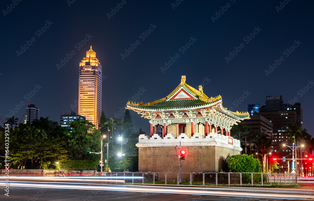 Fototapeta premium The historic Jingfu Gate (East Gate) in a traffic circle at night, with Taipei modern city skyline and traffic light trails in the background, Taiwan