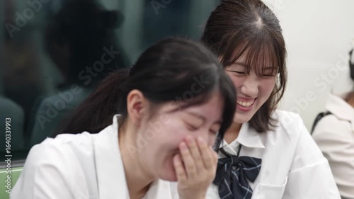 Slow-motion. Japanese high school girls in uniform sit together on a moving train, using smartphones and chatting as the city blurs by beyond window. Shanghai's Pudong New District. China. Spring.