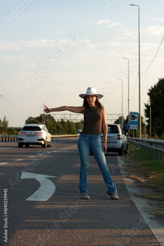 A young beautiful woman in jeans and a hat tries to catch a ride on the highway