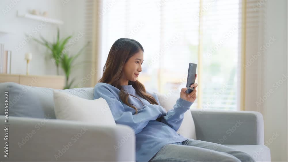 Happy young asian woman relaxing at home. Female smiling and talking chat with friend sitting on sofa and holding mobile smartphone. Girl using video call to close friend Happy young asian woman relaxing at home. Female smiling and talking chat with friend sitting on sofa and holding mobile smartphone. Girl using video call to close friend