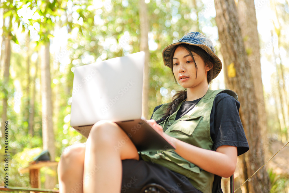 Naklejka premium A woman wearing a green vest and a hat is sitting on the ground and using a laptop. She is looking at the screen