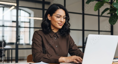 Focused young indian woman wearing glasses and a brown shirt, typing on a laptop at her desk in a contemporary office