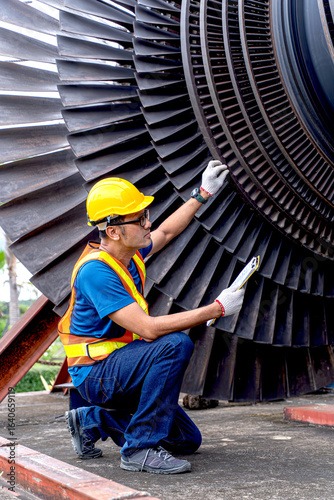 Young Asian worker, power plant engineer in vest and helmet checking of a steam turbine, an essential component in electric power generation on outdoor with clipboard