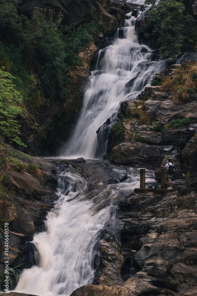 Obraz premium Kuda Ravana Waterfall cascading down rocky terrain in Sri Lanka with tourist admiring the scenery and taking pictures from a wooden platform