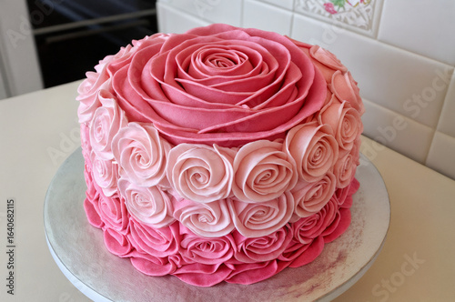 beautifully decorated cake with pink and red rosettes, placed on a round cake board. The background appears to be a kitchen setting with a white backsplash and a stove.