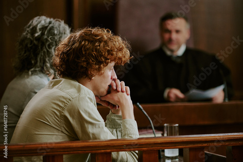 Φωτογραφία Caucasian teenager sitting at witness stand in courtroom, resting chin on claspe