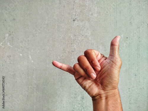 Close-up of a human hand gesturing the 'shaka' sign, also known as the 'hang loose' sign, against a worn, gray wall. The fingers are curled with the thumb and little finger extended.