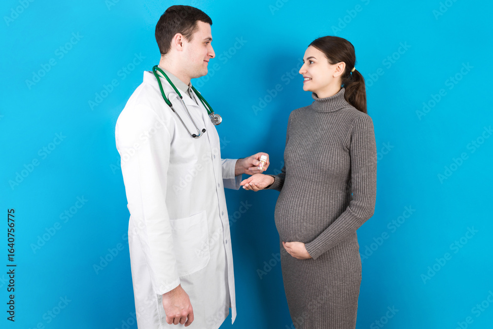 Male gynecologist giving pills to pregnant woman during appointment at colored background. Visit To A Doctor Pregnancy Healthcare And People Concept Male gynecologist giving pills to pregnant woman during appointment at colored background. Visit To A Doctor Pregnancy Healthcare And People Concept