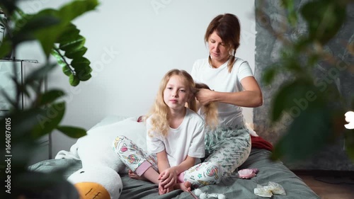 Mother braiding her daughter hair in cozy home setting. Both wearing white T-shirts and colorful pajama pants, sitting on bed with plush toy. Warm family moment of care and bonding. Mothers day