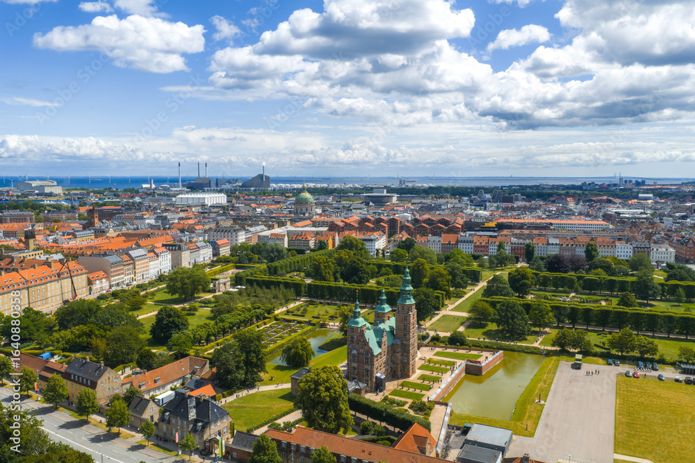 Fototapeta premium Aerial image of Rosenborg Castle with lush gardens, red roofed buildings, Frederik's Church dome, and industrial structures in Copenhagen, Denmark.