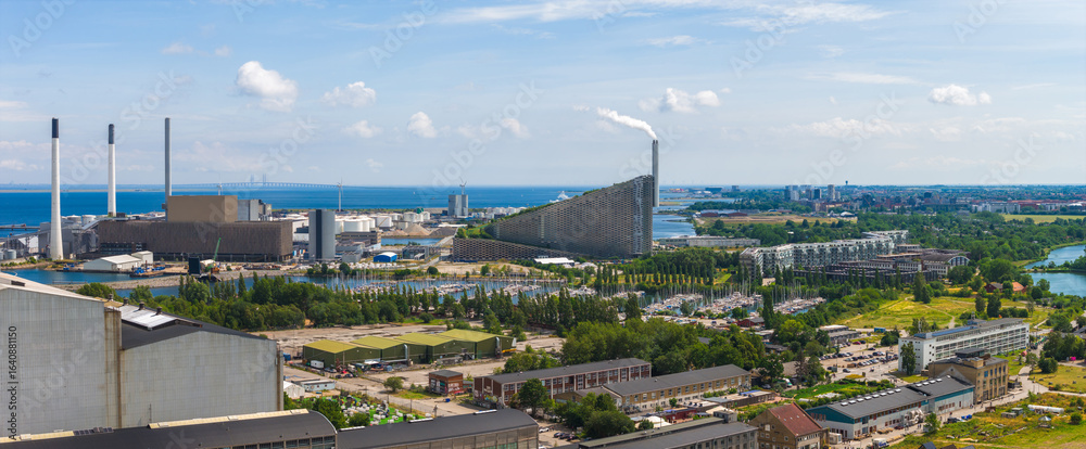 Fototapeta premium Aerial view of Copenhagen, Denmark, showing a waste to energy plant, wind turbines, and the Oresund Bridge over blue waters under a clear sky.