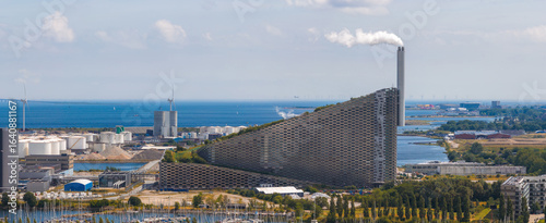Photography Aerial view of Amager Bakke in Copenhagen, Denmark, featuring its sloped green roof, chimney, nearby wind turbines, and the Oresund Strait in the background