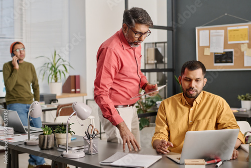 Selective focus shot of Indian men wearing smart casual outfits looking at laptop screen discussing presentation design, their colleague talking on phone in background