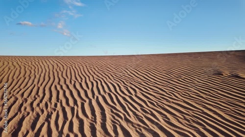 Wind gently flowing across golden sand dunes, forming mesmerizing ripples under expansive blue sky with soft clouds drifting in serene slow motion landscape