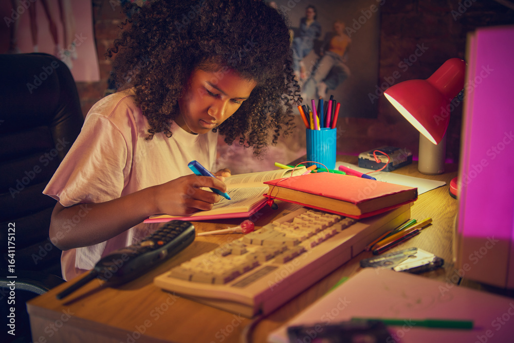Fototapeta premium Teen girl carefully highlights text in open book at wooden desk with vintage computer. Concept of study process, organization, and productivity in a nostalgic workspace.
