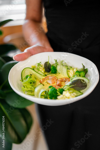 Wallpaper Mural Close-up of a hand holding a bowl of fresh shrimp salad with cucumber, broccoli, lettuce, and diced vegetables. Healthy and colorful meal in a natural setting. Torontodigital.ca