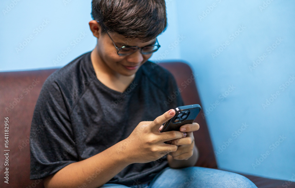 Fototapeta premium Asian guy teenager sitting on sofa at home relax using mobile phone app typing text browsing message, gen z checking social media on webpage, happiness lifestyle digital communication concepts