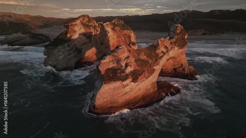 Aerial view of Wharariki Arch rocks sculpted by the ocean, the sun paints the rock formations in warm shades of orange against the dark blue sea, Puponga, New Zealand.