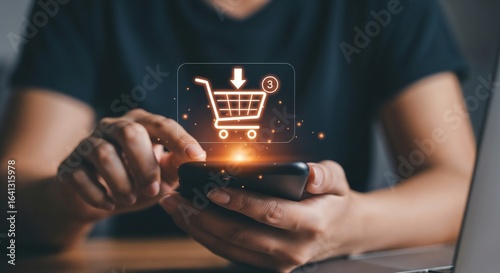 A woman's hands add an item to her online shopping cart using a smartphone. A luminous orange shopping cart hologram is displayed above the device. A laptop is visible in the background.