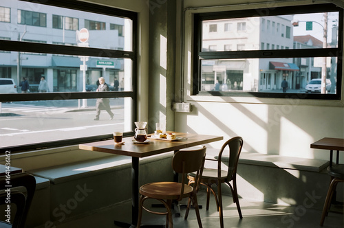A quiet and minimalist cafe interior features a wooden table with a view of the street outside. Sunlight streams in, creating a peaceful and contemplative mood.