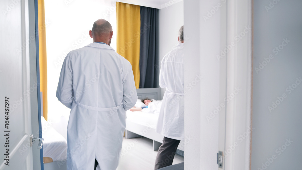Fototapeta premium Two doctors enter the patient's room, view from the back. Two men in white coats are standing in front of a sick man's bed. Morning rounds of patients at the clinic