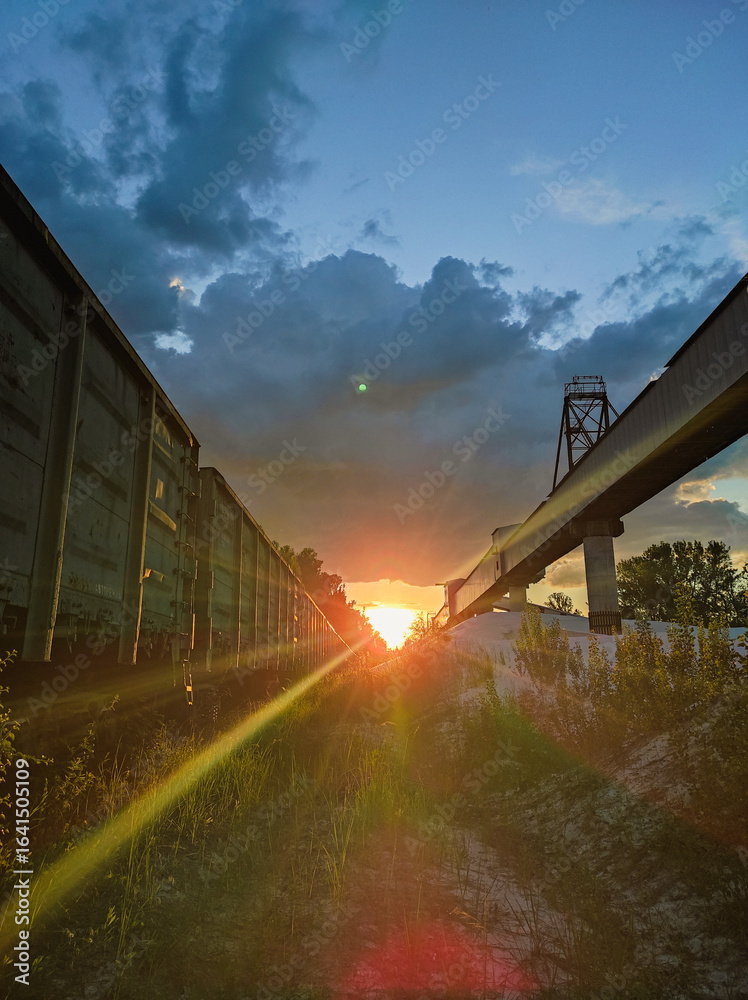 Fototapeta premium A beautiful sunset with a prominent sun flare illuminates the space between a long green freight train and a high industrial conveyor structure, surrounded by green grass and a dramatic, cloudy sky.